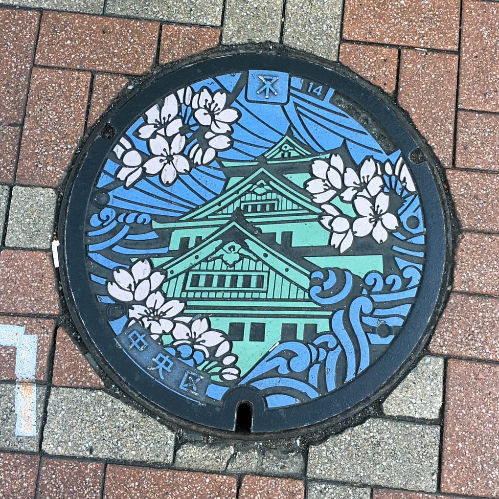 A colorful manhole cover in Osaka. Osaka castle is depicted in green, surrounded by pink cherry blossoms and blue water