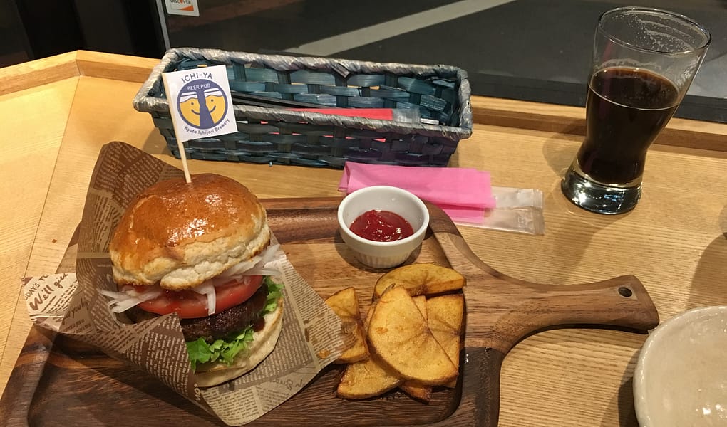A small hamburger on a wooden cutting board sits next to a stout beer