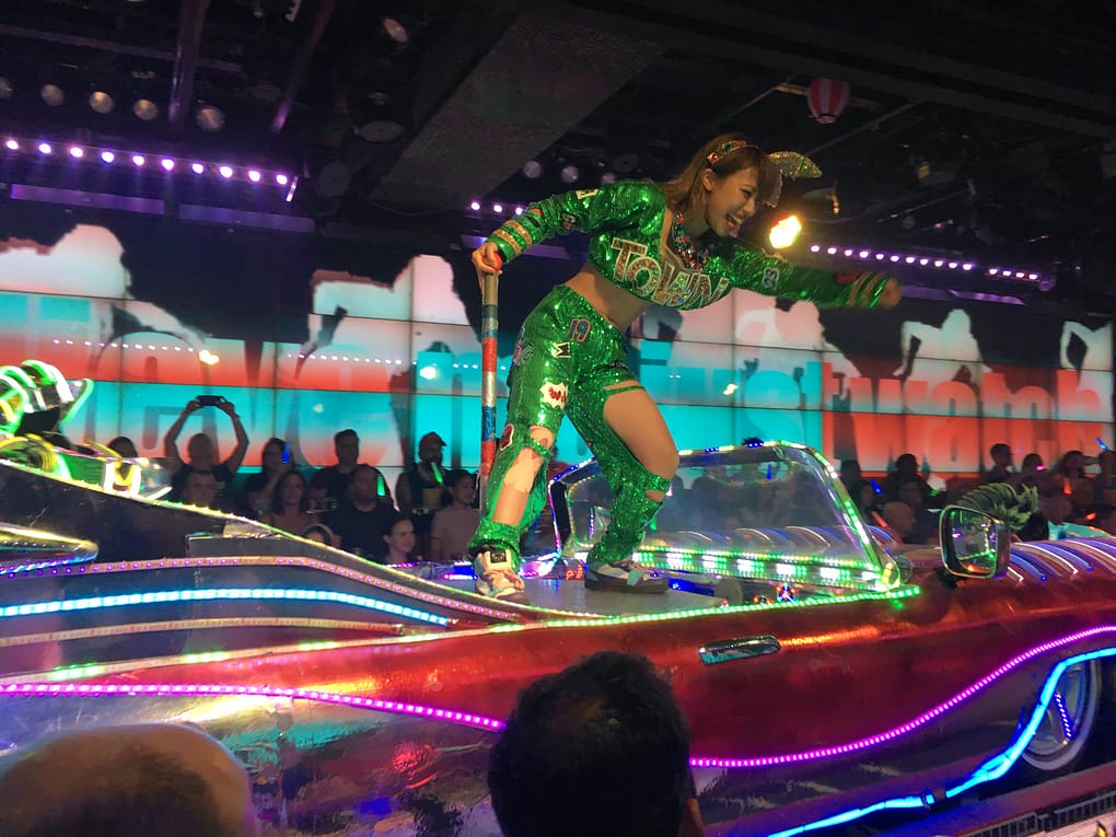 A performer dressed in green stands on top of a convertible with neon accents