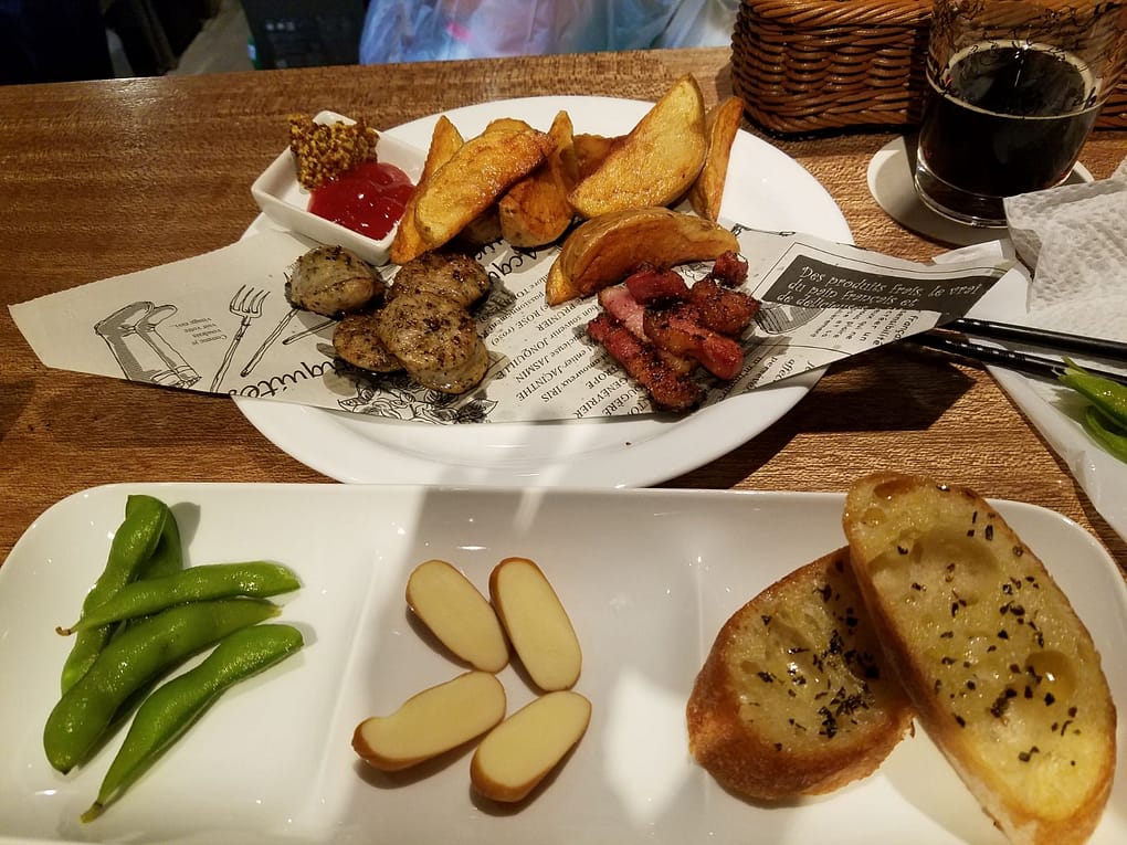 Two plates of food sit on a wooden bar. The round plate in the back has sausage, bacon and fries. The rectangular plate in the foreground has edamame, cheese and bread.