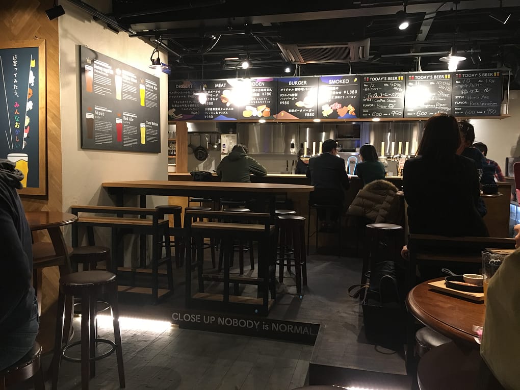 The inside of Beer Pub Ichi ya in Kyoto. There are high top tables, with customers sitting at the bar. chalkboards on the back wall provide a list of beers for sale.