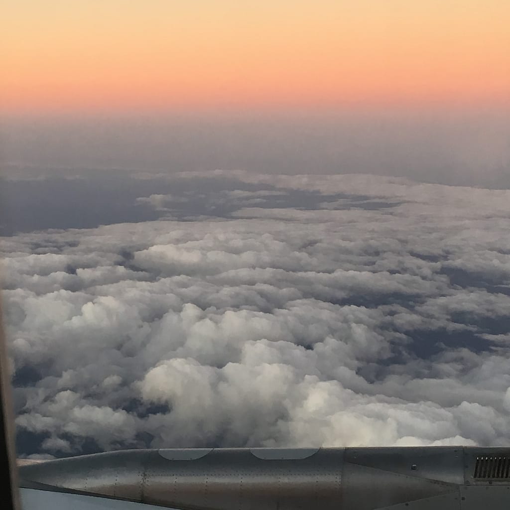 A sky view from the window of an airplane. The sun is setting in the background with clouds in the foreground