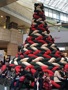 A tall red, white and green Christmas tree made out of plush material. Visitors sit for a rest at the base.