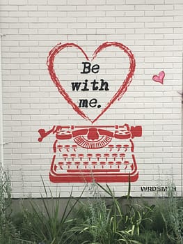 a white brick wall with a red spray painted typewriter. Above the typewriter is a heart that says "Be with me"