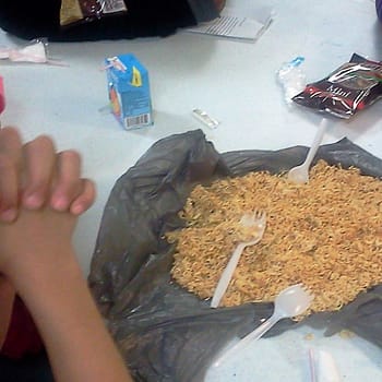 A child's hands are clasped in prayer. A plastic bag of dried ramen noodles and plastic cutlery sits on a table.