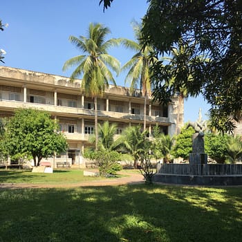 The outside courtyard of Tuol Sleng. There are palm trees in the yard and a concrete building in the background