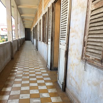 Checkboard tile floors lead the way down an outside breezeway. Wooden shutters and concrete walls mark the rooms of Tuol Sleng