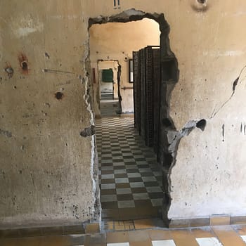 A doorway is broken into a concrete wall. Through the wall one can see the wooden doors of cells in Tuol Sleng