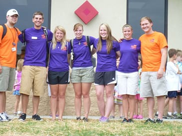 Seven adults line up in a row with arms behind each others backs. Two males are on the left, four females in the middle, and one male on the right. The two males on the end are wearing orange shirts. The five in the middle are wearing purple shirts