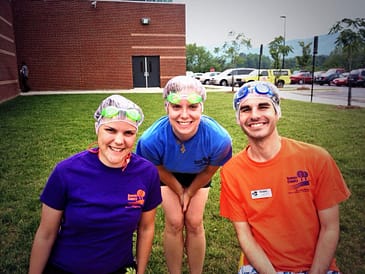 Two females and one male wear swimming goggles and shower caps. The female and male in the front are sitting and one female in the back in standing