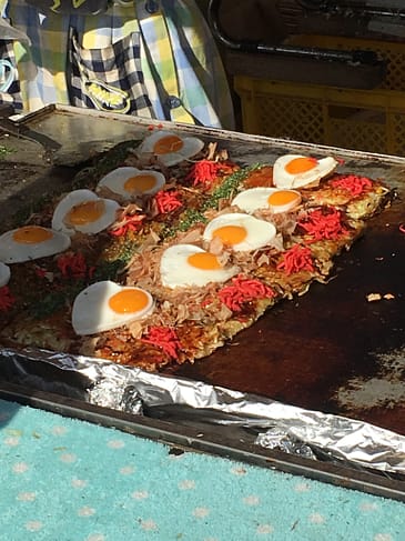 A street vendor cooks food on a skillet. There are two rows of food, and each serving has a pancake type base, meat, pickled ginger, and an egg