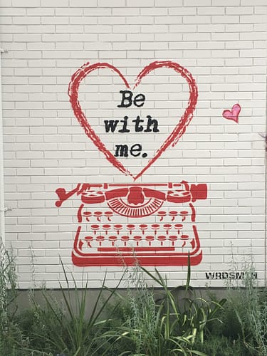 spray painted street art on a white brick wall. A red typewriter with a red heart that says "be with me" in black text