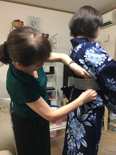 An older woman stands behind a younger woman. She is fastening a wide obi-ita around her waist.