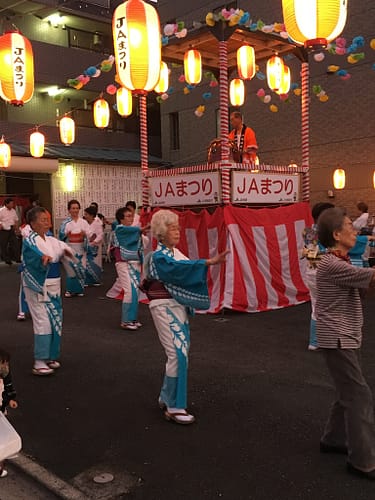 There's a dj in an elevated booth, surrounded by yellow-white lanterns. Dancers are below, many wearing the same style yukata.