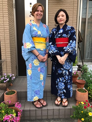The woman on the left wears a light blue yukata with a yellow obi. On the right is a woman dressed in a dark blue yukata with a red obi.