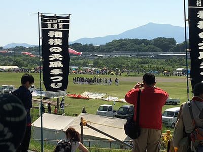 Image shows mountains in the background. Middle ground has a group of darkly dressed people clustered around the rope that serves as the kite string. Foreground shows festival flags and the backs of other spectators