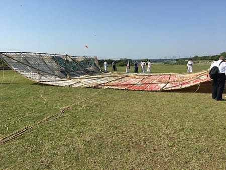 Image shows a large kite laying face down on the ground. The kite is constructed with bamboo sticks and thin paper. People stand around the back