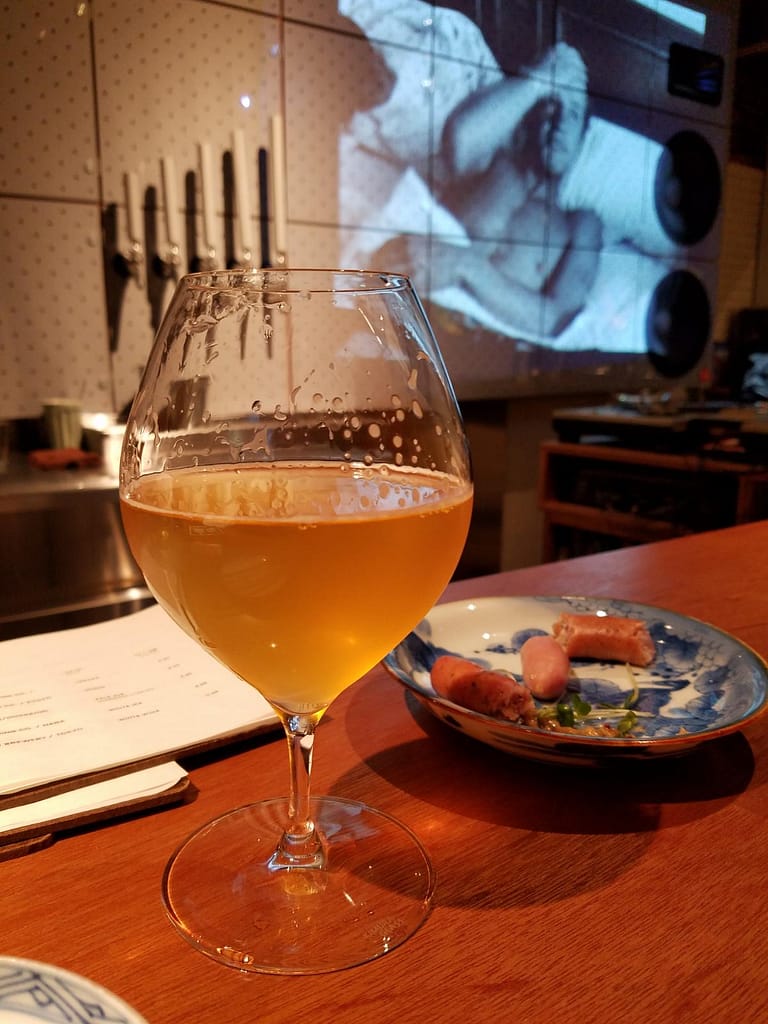 A light colored beer in a rounded glass next to a small plate of sausage