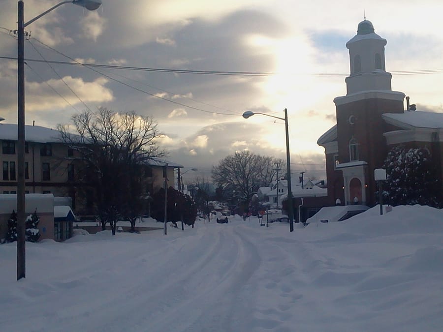 A snow covered street with a church on the right and the sun setting in the background