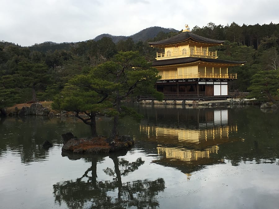 Kinkaku-ji, a temple covered in gold leaf, sits on the banks of a pond. The image of the temple is reflected in the pond's still waters