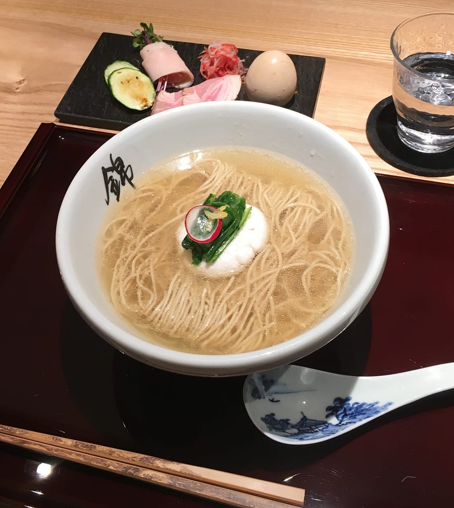 A bowl of ramen topped with an egg sits on a wooden tray. An appetizer of red snapper dumplings is in the background